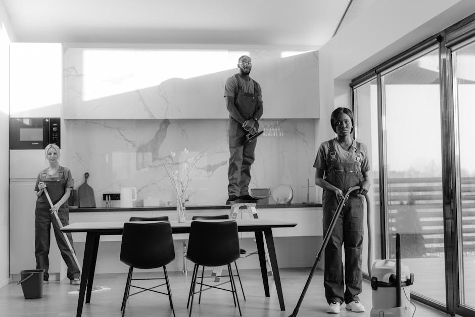 A black and white photograph of a modern kitchen with a minimalist design, featuring a large marble backsplash, a sleek white countertop, and a wooden dining table surrounded by black chairs. Three cleaning professionals wearing uniform overalls are present: one standing on a ladder cleaning the upper part of the backsplash, another mopping the tiled floor near the window, and the third holding a cloth, ready to clean the surface. The room is well-lit with natural light coming through large sliding glass doors that lead to an outdoor balcony. Visible cleaning tools include a mop, a bucket, and a vacuum cleaner. The scene emphasizes surface cleaning, deep cleaning, and sanitisation processes carried out by Pimlico Cleaner in a domestic setting, ensuring high standards of hygiene and cleanliness.