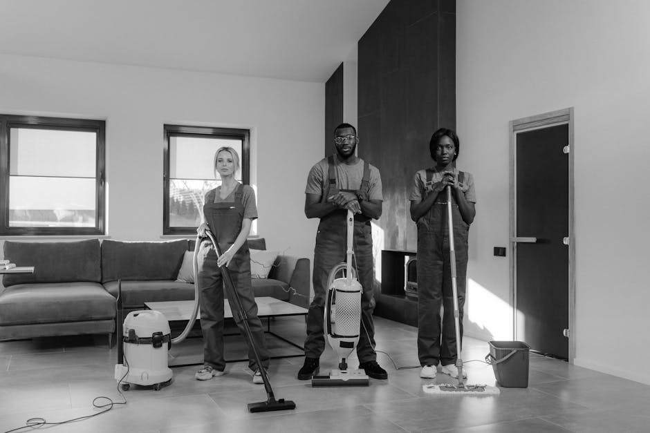 A black and white photograph of a modern kitchen with a minimalist design, featuring a large marble backsplash, a sleek white countertop, and a wooden dining table surrounded by black chairs. Three cleaning professionals wearing uniform overalls are present: one standing on a ladder cleaning the upper part of the backsplash, another mopping the tiled floor near the window, and the third holding a cloth, ready to clean the surface. The room is well-lit with natural light coming through large sliding glass doors that lead to an outdoor balcony. Visible cleaning tools include a mop, a bucket, and a vacuum cleaner. The scene emphasizes surface cleaning, deep cleaning, and sanitisation processes carried out by Pimlico Cleaner in a domestic setting, ensuring high standards of hygiene and cleanliness.