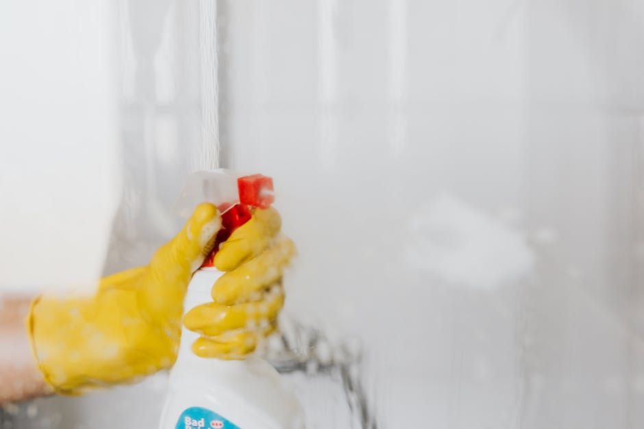Close-up of a person's hand wearing a yellow rubber glove, holding a white spray bottle with a red nozzle, actively wiping a vertical, glossy white tiled surface in a modern kitchen or bathroom. The surface appears clean and shiny, reflecting light from the surroundings, with some visible water or cleaning solution spray. The background is blurred, featuring additional white tiles and soft natural lighting. This scene illustrates surface cleaning and sanitisation practices aligned with domestic cleaning services provided by Pimlico Cleaner, emphasizing hygiene and meticulous upkeep of tiled surfaces.