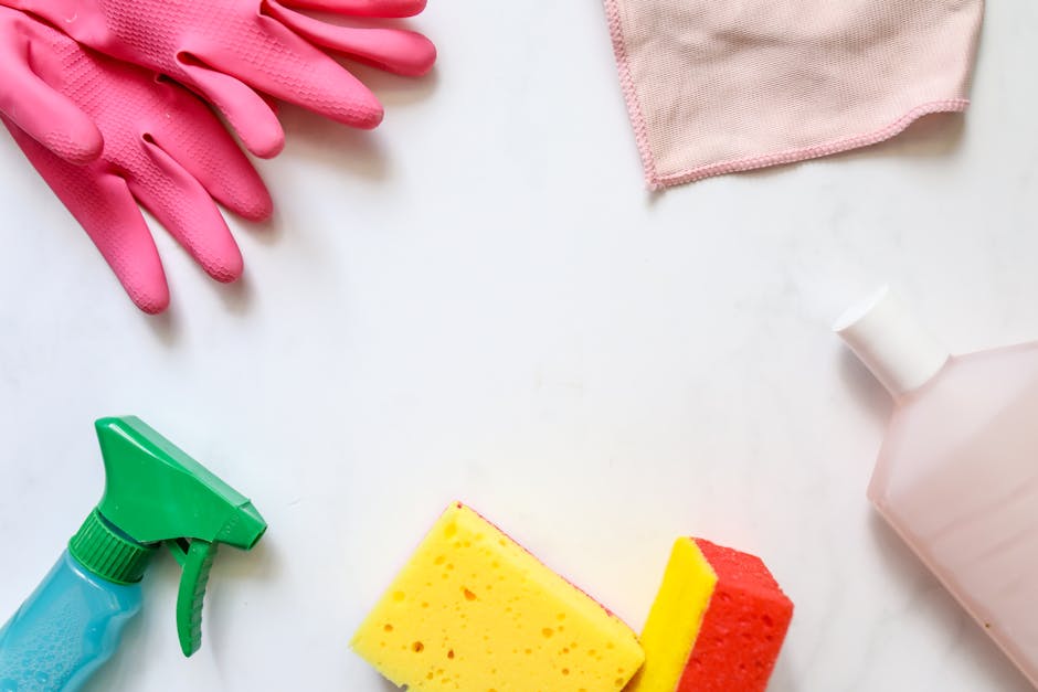 A flat lay arrangement of cleaning tools and supplies on a white surface, including a pair of pink rubber gloves in the top left corner, a pink cloth in the top right corner, a green spray bottle with a blue liquid solution in the bottom left, a yellow and red cleaning sponge in the bottom center, and a white plastic bottle in the bottom right, all situated in a well-lit environment indicating surface cleaning and maintenance as part of professional domestic cleaning services promoted by Pimlico Cleaner near Tate Britain, Pimlico.