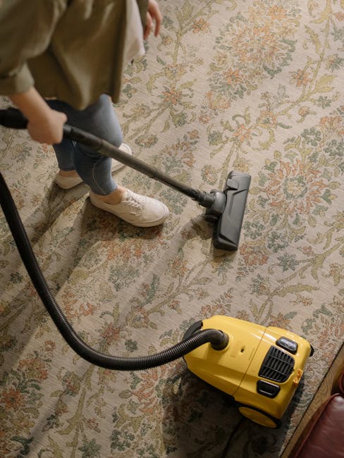 A woman performing surface cleaning and deep cleaning in a living room, using a yellow vacuum cleaner with a hose attachment to clean a patterned carpet. She is dressed in casual clothing, including a