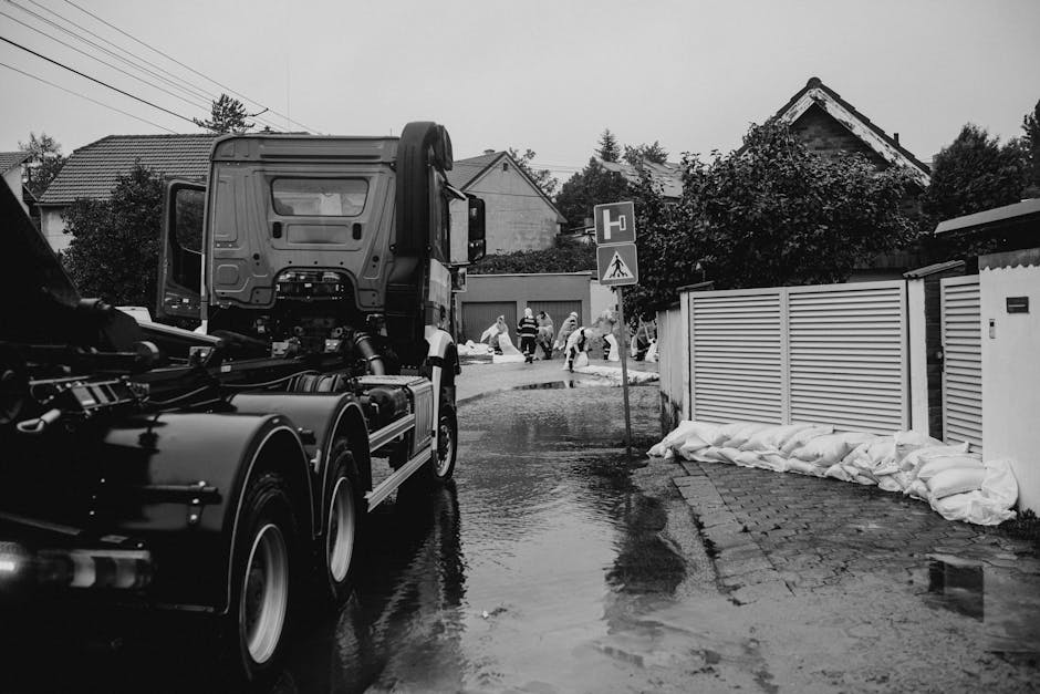 A residential street scene showing a large truck actively engaged in emergency flood response, with workers in high-visibility clothing standing on the wet pavement. The road is flooded with puddles r