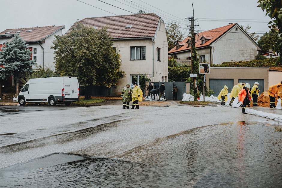 A residential street scene showing a large truck actively engaged in emergency flood response, with workers in high-visibility clothing standing on the wet pavement. The road is flooded with puddles r