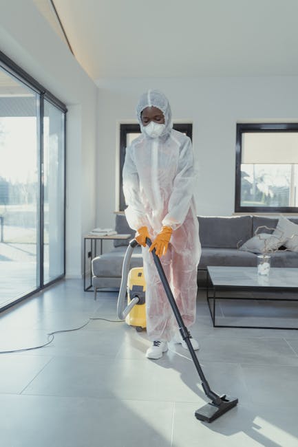 A person dressed in white protective clothing, including a full-body suit, gloves, and shoe covers, is performing surface cleaning or sanitisation on a wall-mounted flat-screen television in a modern living room. The room features a white brick wall, a black armchair with a grey throw draped over it, and a large green potted plant near a glass door that leads to an outside balcony. The wooden floor appears clean, and a black coffee table and framed pictures are visible in the background. The lighting is natural, coming from the nearby balcony, and the scene emphasizes thorough cleaning and maintenance to ensure hygiene and sanitisation, as offered by Pimlico Cleaner with their mould removal solutions for basement flats in Pimlico.
