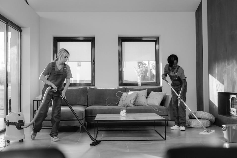 Three professional cleaning staff in an uncluttered living room preparing for surface cleaning and deep cleaning tasks. The room features a large L-shaped sofa with fabric upholstery, a rectangular co