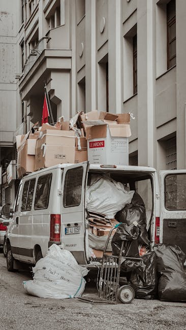 A white van parked on a city street adjacent to a multi-storey building with beige stone façade and rectangular windows. The van's rear doors are open, revealing various large black and white garbage
