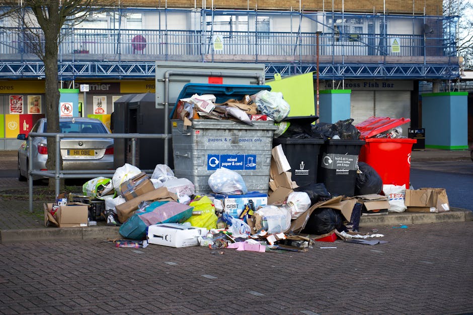 A public waste disposal area in front of a commercial building, featuring a large metal recycling bin filled with mixed paper and cardboard, with its lid open and various papers and packaging overflowing onto the surrounding pavement. Several black and red rubbish bags are placed next to the bin, some partially torn open, spilling waste onto the brick-paved ground. Nearby, a silver car is parked beside a metal railing, with a tree and shopfronts in the background. The scene appears untidy with scattered packaging and trash, highlighting the importance of proper waste management and cleaning practices for maintaining cleanliness in accordance with Westminster Council rules, as promoted by Pimlico Cleaner.