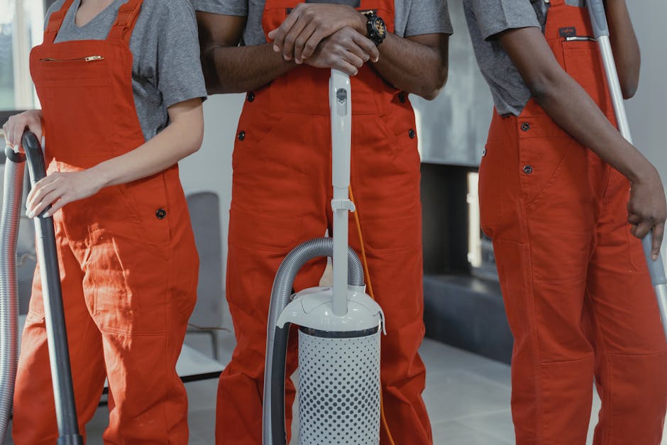 Close-up of three professional cleaners dressed in grey shirts and bright orange uniforms, each holding a vacuum cleaner or cleaning tool, performing surface cleaning in an indoor setting. The floor features large ceramic tiles with a clean, shiny appearance, reflecting good maintenance. One cleaner holds a handheld vacuum connected to a main unit, while another holds a wand attachment. The background shows a modern, well-lit space with neutral-colored walls and minimal furniture, emphasizing hygiene and deep cleaning procedures that Pimlico Cleaner provides for residential or commercial spaces. The scene highlights the importance of sanitisation and thorough surface maintenance aligned with HMO rules in Pimlico.