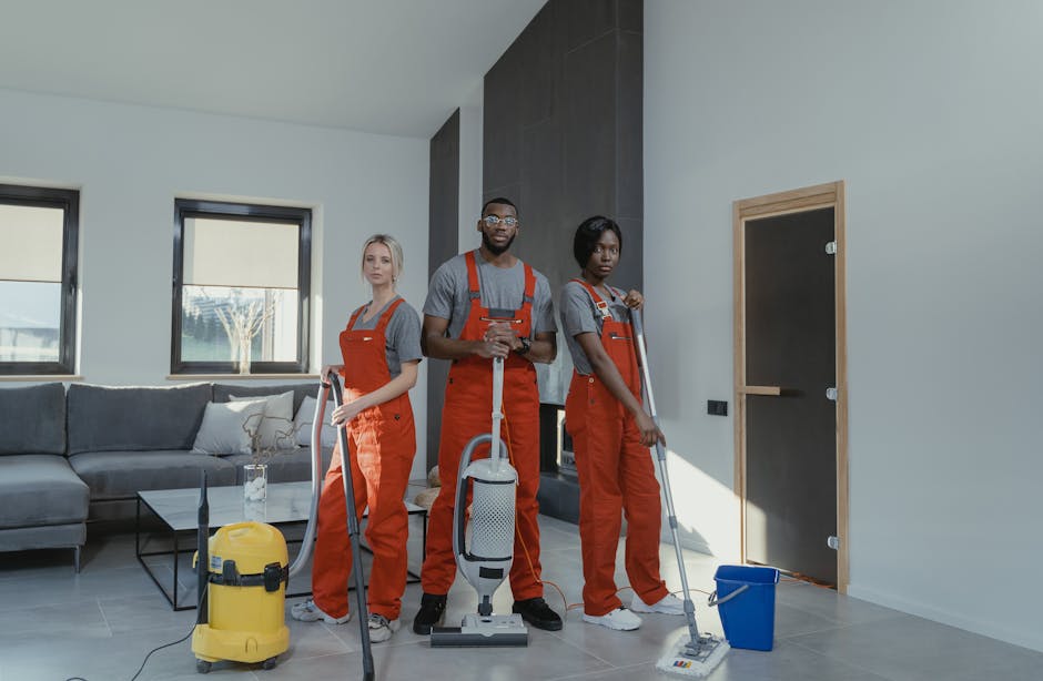 Close-up of three professional cleaners dressed in grey shirts and bright orange uniforms, each holding a vacuum cleaner or cleaning tool, performing surface cleaning in an indoor setting. The floor features large ceramic tiles with a clean, shiny appearance, reflecting good maintenance. One cleaner holds a handheld vacuum connected to a main unit, while another holds a wand attachment. The background shows a modern, well-lit space with neutral-colored walls and minimal furniture, emphasizing hygiene and deep cleaning procedures that Pimlico Cleaner provides for residential or commercial spaces. The scene highlights the importance of sanitisation and thorough surface maintenance aligned with HMO rules in Pimlico.